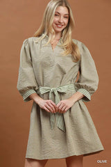 Woman wearing Umgee olive textured contrast trim tie waist mini dress with striped belt and puff sleeves.